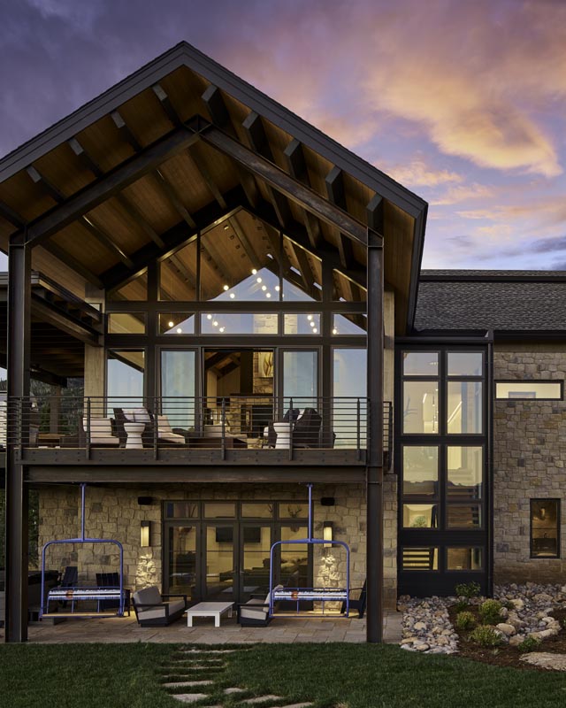 A modern two-story house with large glass windows and a stone facade under a vibrant sunset sky. the home features a balcony with illuminated interiors and artistic lighting.