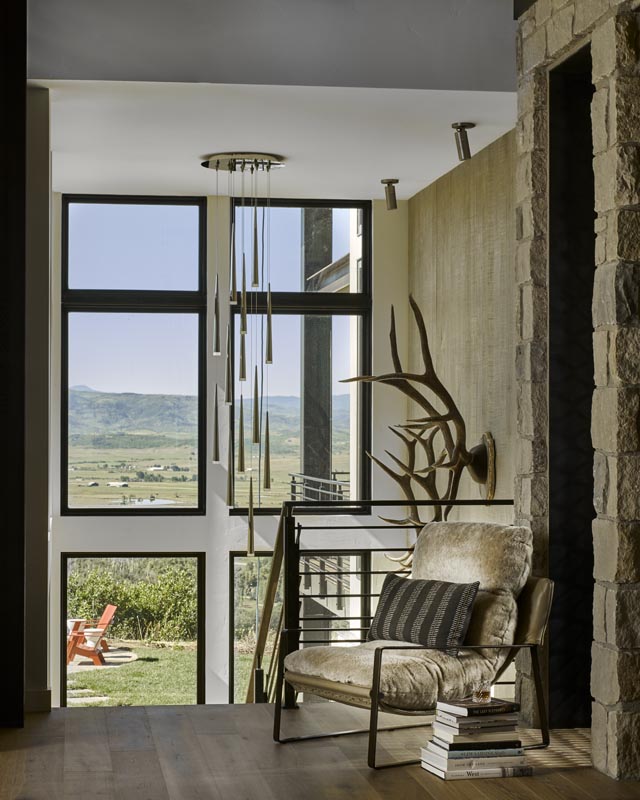A cozy reading nook with a plush chair, stacked books, and a floor lamp, beside large windows showing a scenic view of a green valley and mountains, complemented by stylish, minimalist decor.