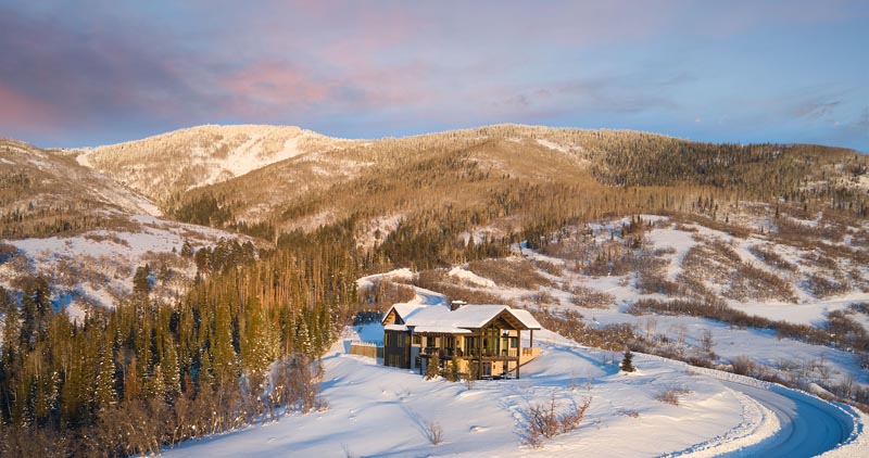 A large house with a dark roof sits surrounded by snow-covered hills and forests during sunset, with pink clouds in the sky above.