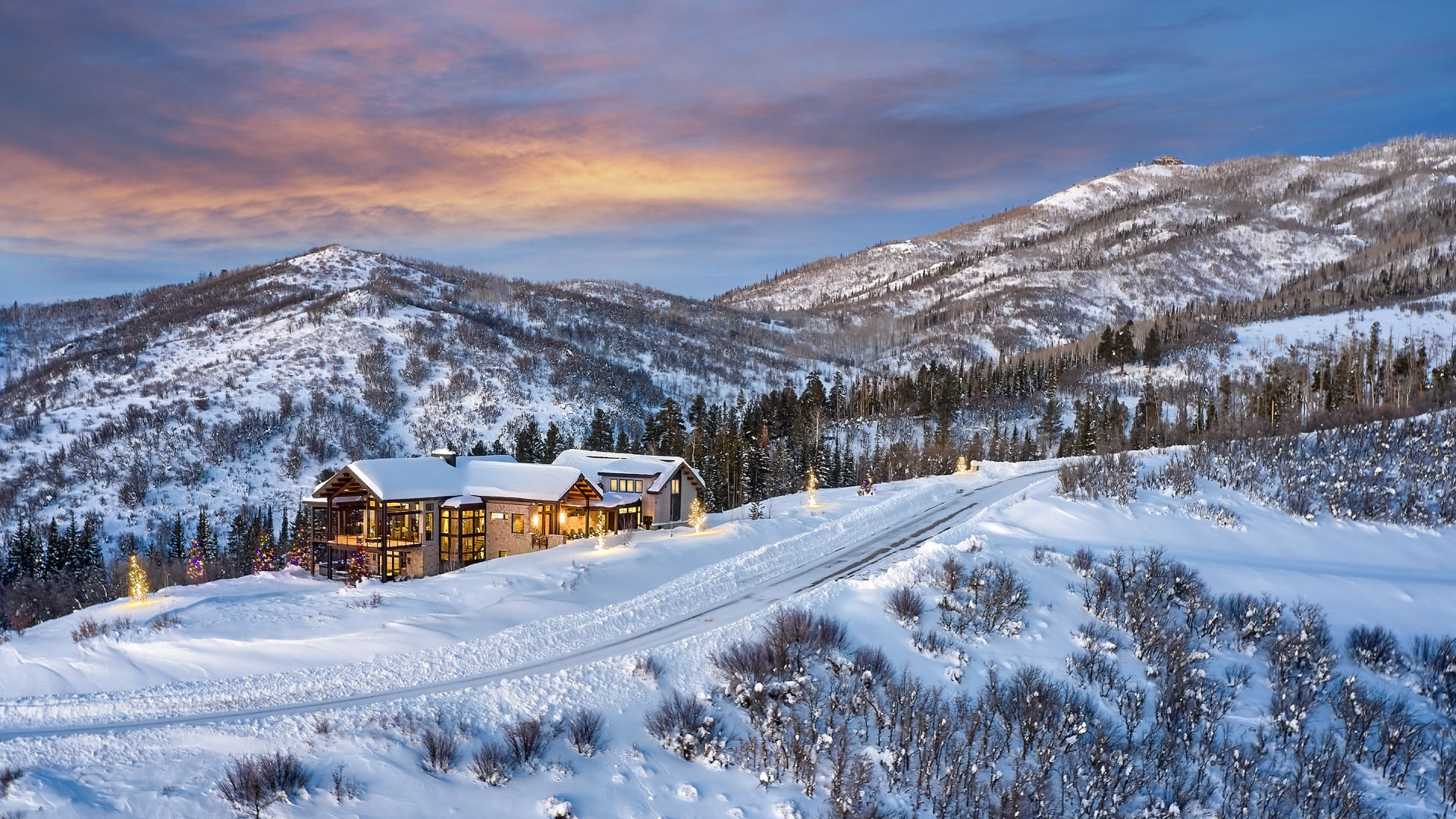 A winter landscape at dusk featuring a snow-covered mountain and a cozy, illuminated lodge designed and built surrounded by trees, under a vibrant orange and blue sky.