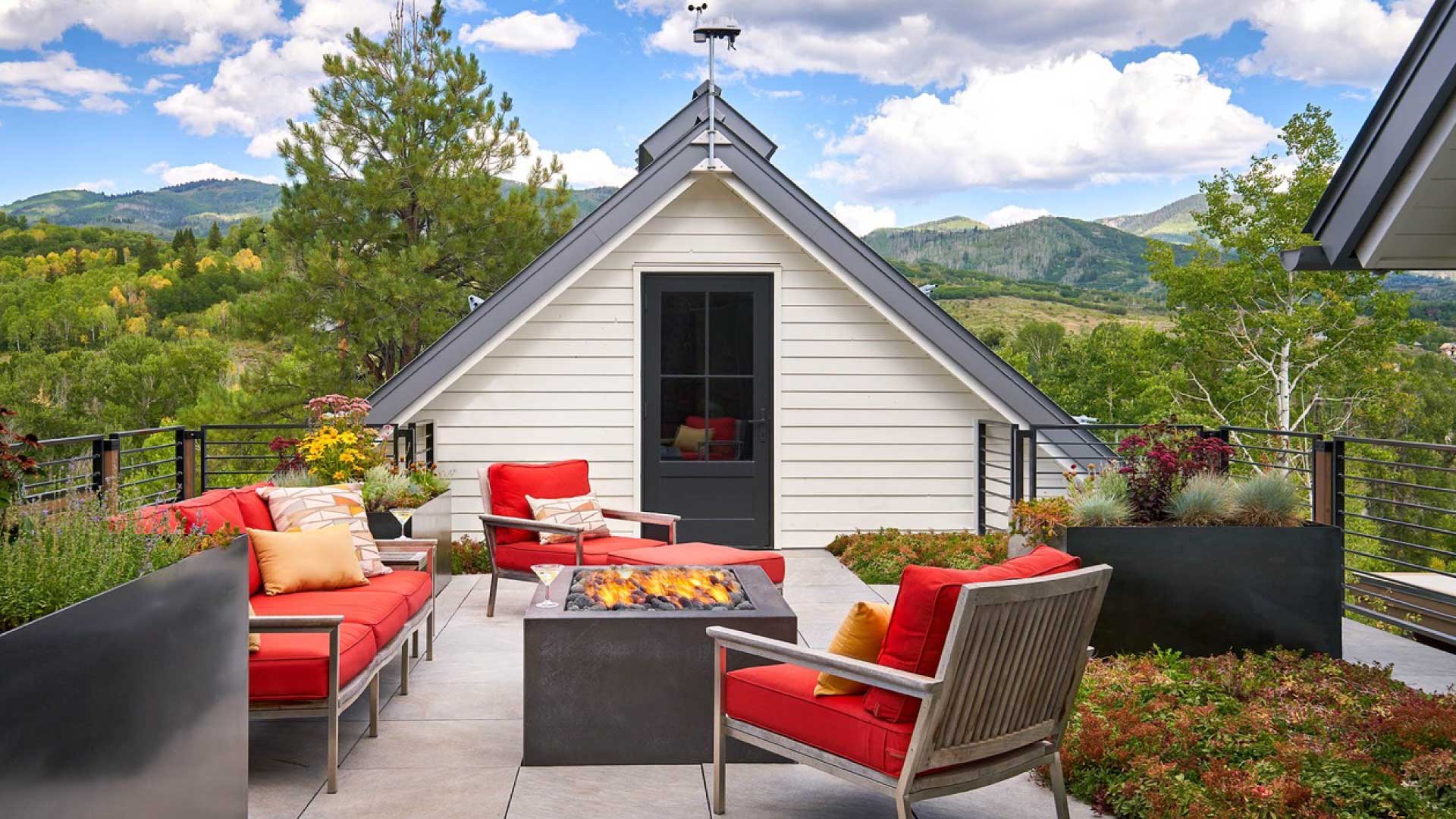 A cozy outdoor terrace with a fire pit surrounded by red and grey modern furniture, offering a view of green hills and a charming white a-frame house.