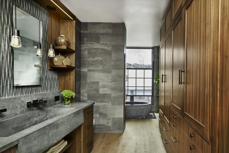 Modern bathroom featuring wooden cabinetry, gray stone sink and wall, mirror with sconces, and a view to an outdoor area through a large window.