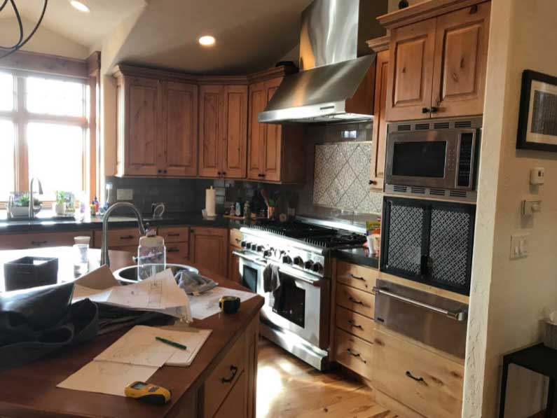 GB-design-steamboat-springs-colorado-Renovations-Lakeside-Living-Reimagined-1b A spacious kitchen with wooden cabinetry, stainless steel appliances including a range hood and stove, and a well-lit window by the sink. there are papers and items on the counter.