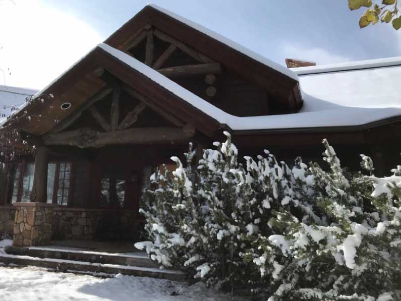 GB-design-steamboat-springs-colorado-Renovations-Lakeside-Living-Reimagined-2b A cozy snow-covered cabin with a sloped roof and exposed wooden beams, surrounded by snow-dusted bushes under a cloudy sky.