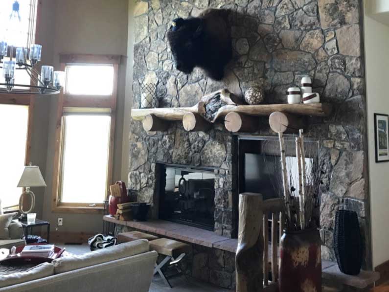 GB-design-steamboat-springs-colorado-Renovations-Lakeside-Living-Reimagined-3b A cozy living room with a stone fireplace, a mounted bison head above the mantel, and rustic decor including a wooden bench and various decorative items.