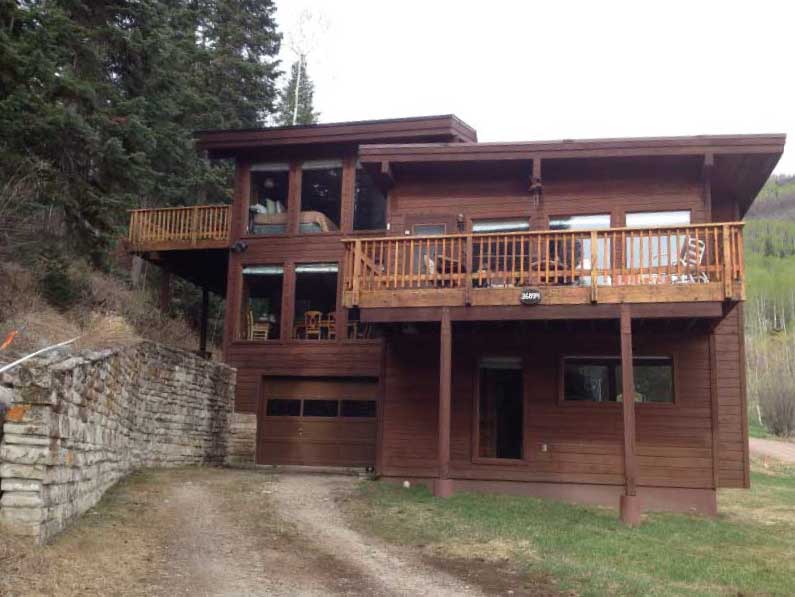 GB-design-steamboat-springs-colorado-Renovations-Tree-Haus-Transformation-1b A two-story wooden house with balconies nestled in a leafy hillside area. the house has a garage at the ground level and is surrounded by light green foliage.