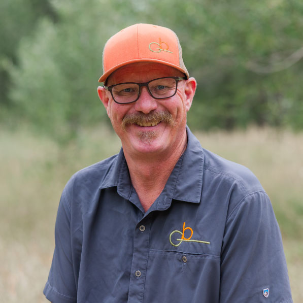 GB-design-steamboat-springs-colorado-about-partners-BLarson A smiling man with glasses, a mustache, and an orange cap, wearing a gray shirt with a yellow embroidered team logo, stands outdoors in a natural, green setting.