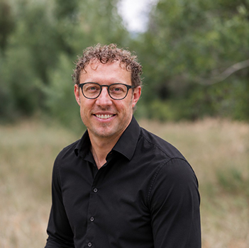2025_0729_GBDesign-20 CFletcher BETTER 350x350 A man with curly hair and glasses wearing a black button-down shirt, smiling outdoors.
