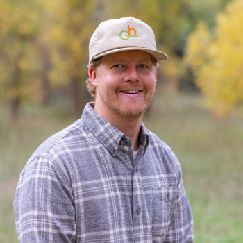 Bobby-Henson A man wearing a plaid shirt and a baseball cap with a logo smiles outdoors.